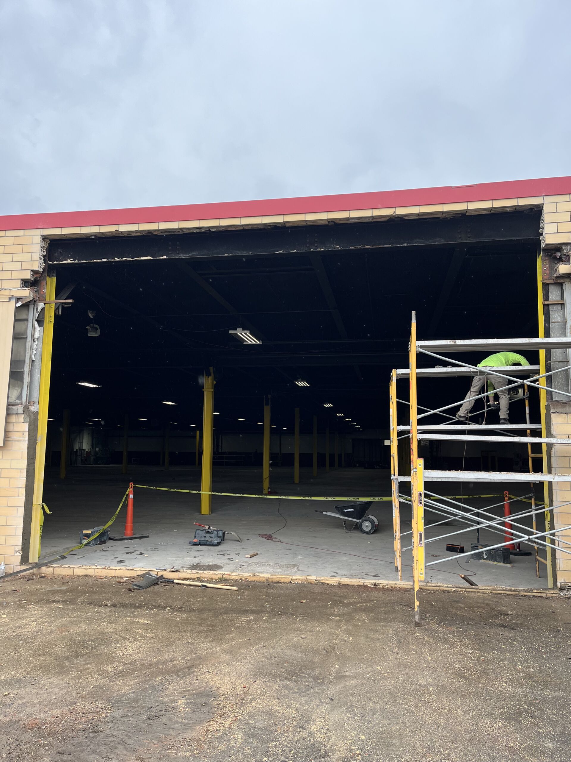 Open warehouse bay under construction with yellow vertical supports and scaffolding on the right; caution tape on the floor and construction debris outside.