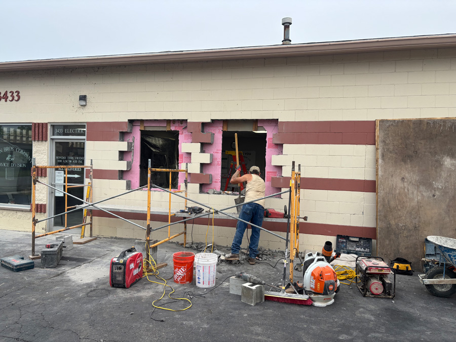 Construction worker at a storefront renovation chipping away at the exterior wall with pink insulation exposed and scaffolding set up outside.