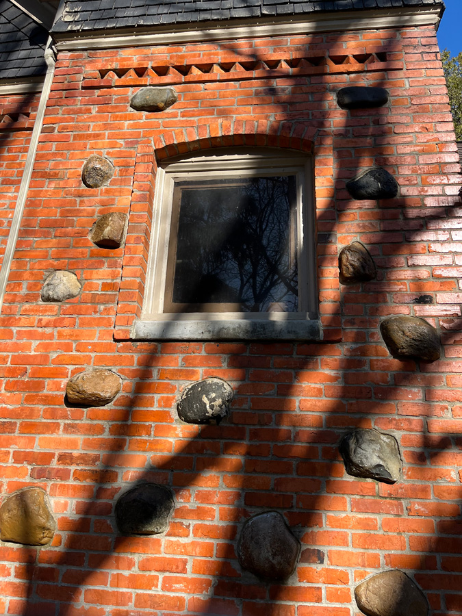 Red brick wall with scattered embedded stones and a small window under an arched brick lintel, with shadows across the surface.