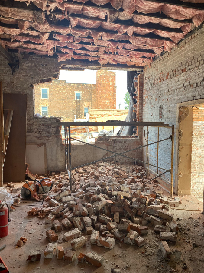Interior of a partially demolished room with a brick wall opening to the outside, rubble and bricks on the floor, and scaffolding in the center.
