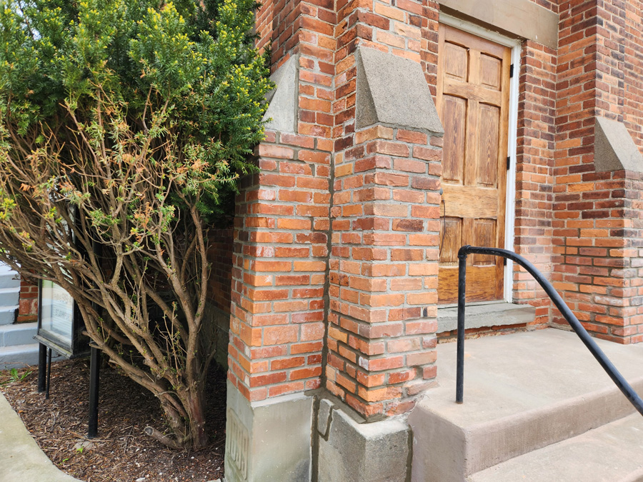 Brick building entrance with a wooden door, concrete steps, and a black handrail; a large shrub grows along the left side near the sidewalk.