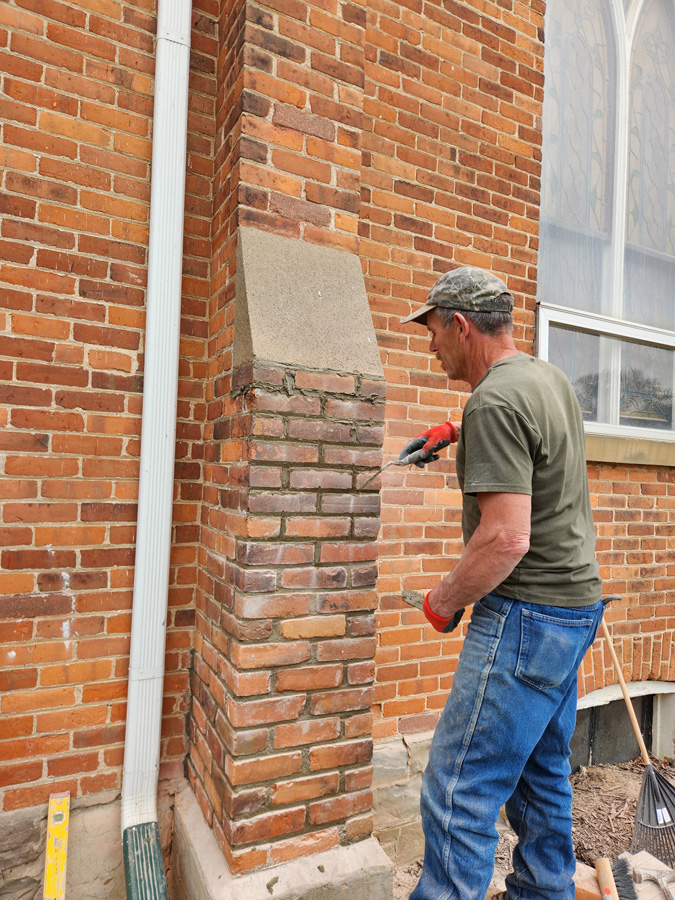 Mason in a green shirt and camouflage cap chisels bricks on an exterior brick column beside a window.