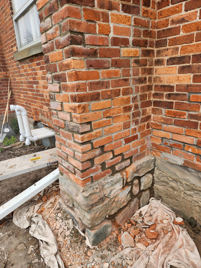 Corner of a brick building showing an exposed stone foundation, with debris and construction tarps nearby.