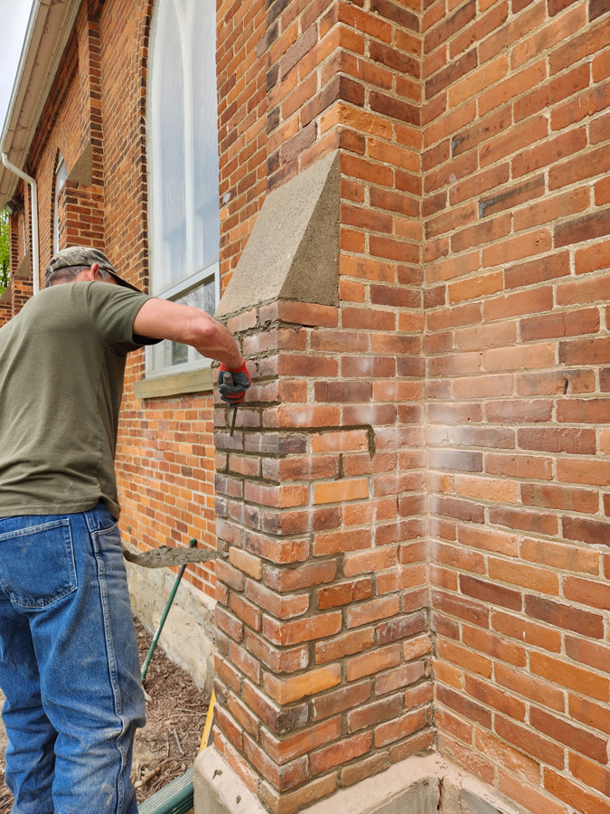 Man in a green shirt repointing an exterior brick wall with a chisel and hammer occupantly outside a house.