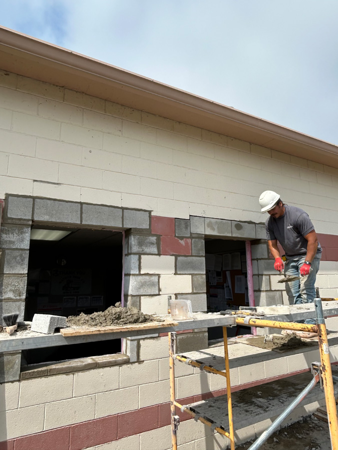 Construction worker wearing a white hard hat and red gloves stands on scaffolding, laying concrete blocks to build a wall on an exterior building.