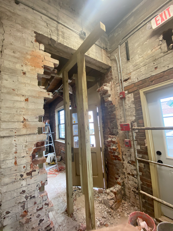 Interior under renovation: exposed brick walls with damaged sections, wooden stud framing, a doorway, debris on the floor, a ladder, and an exit sign nearby.