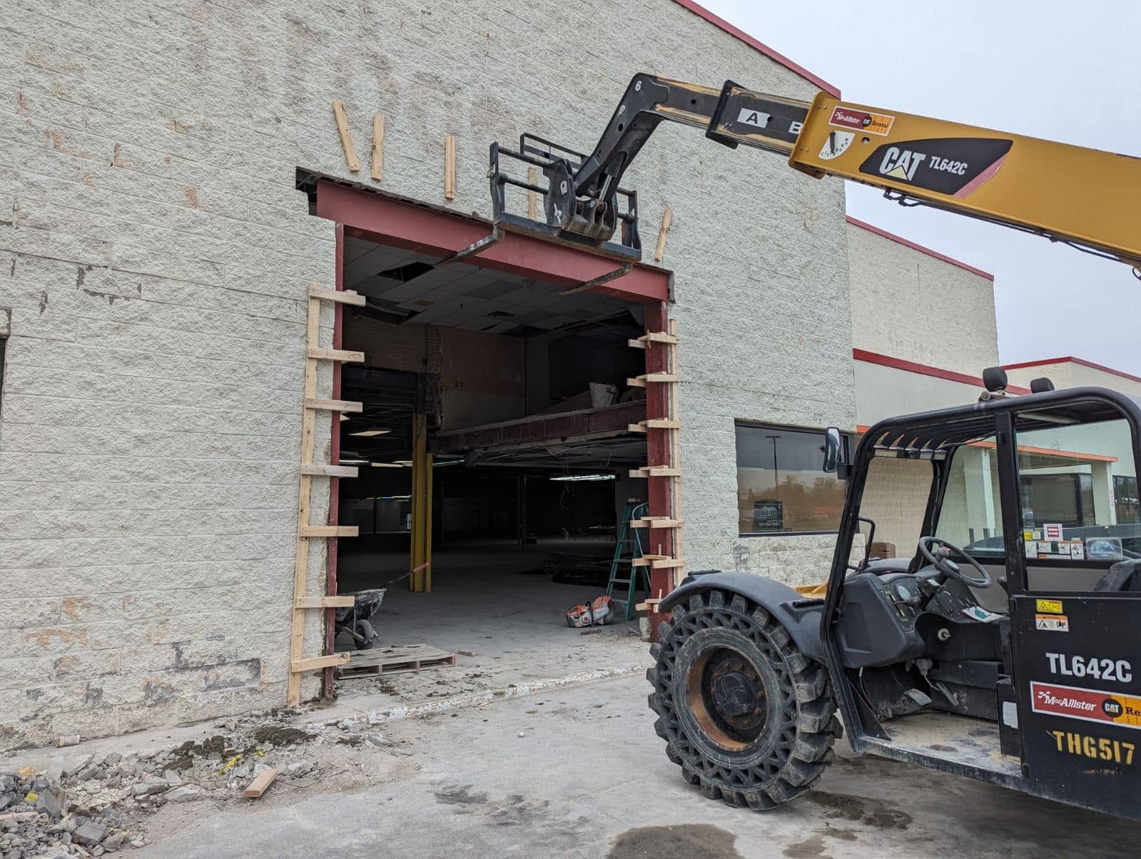 Construction site: a storefront opening being enlarged with wooden framing and a yellow CAT excavator arm overhead, work debris on the ground.