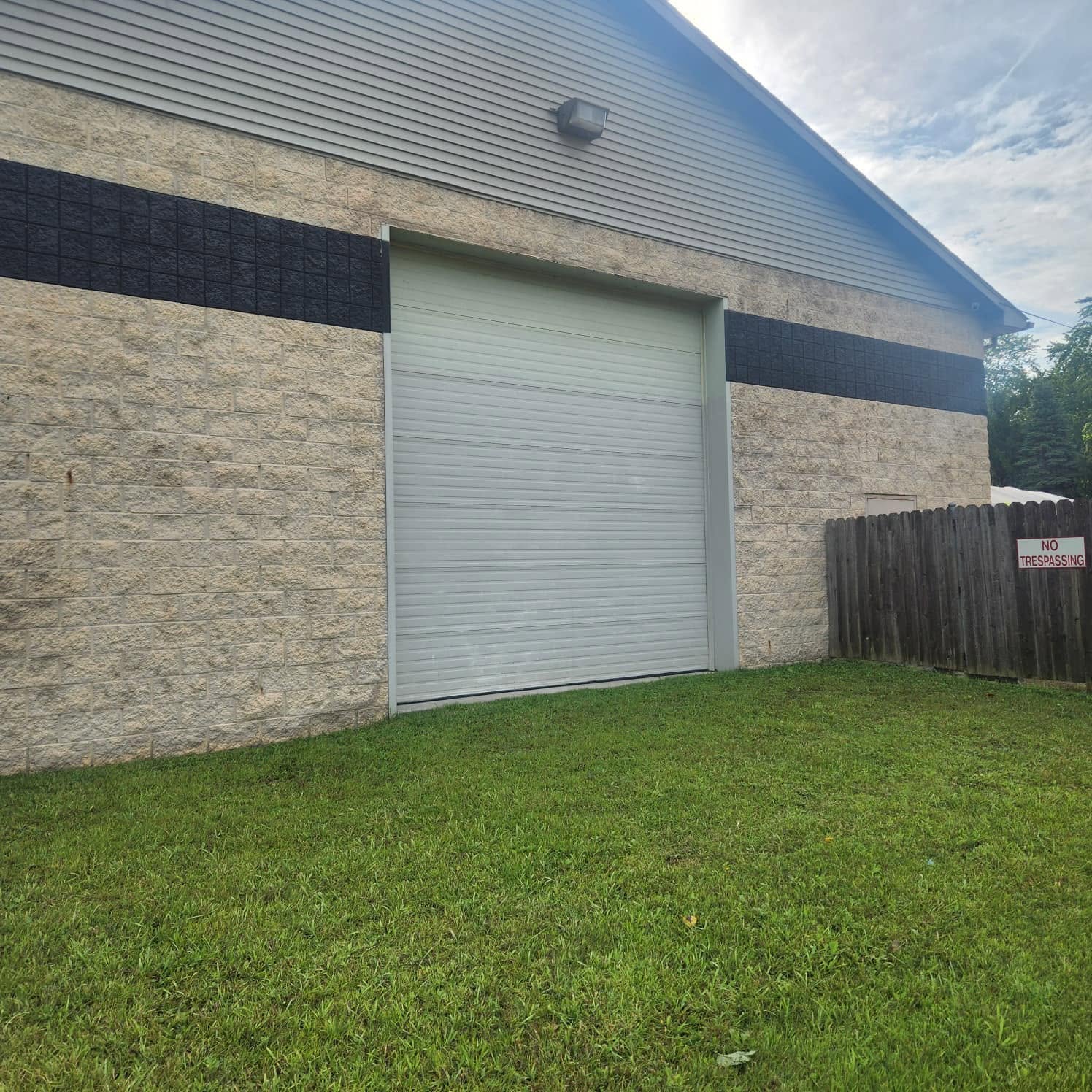 Industrial brick building with a large gray roll-up door; beige walls with a black horizontal strip and a wooden fence nearby with a 'No Trespassing' sign on a sunny day.