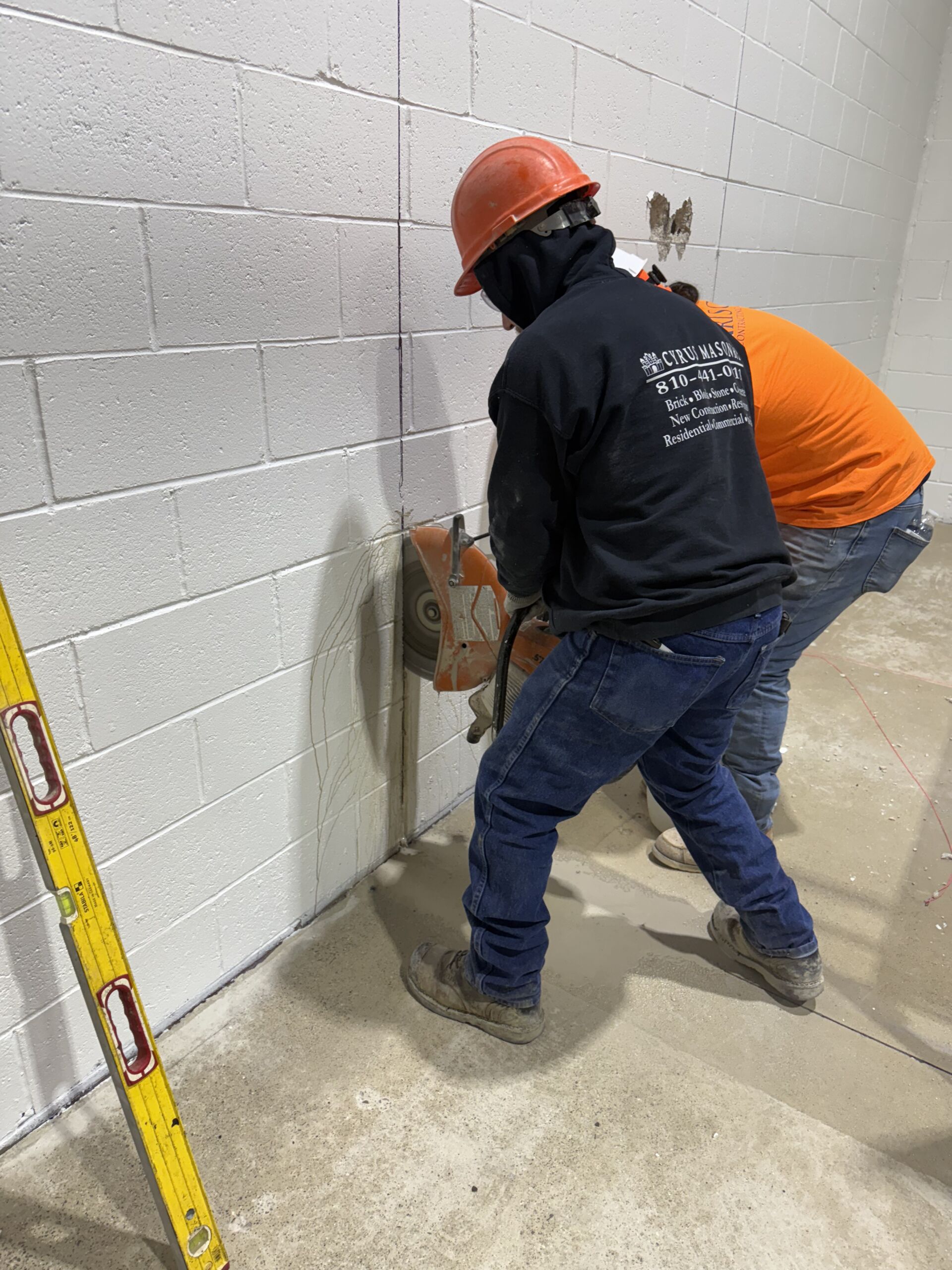 Two construction workers wearing hard hats and hoodies cut into a concrete block wall with a handheld grinder; a yellow level rests on the floor nearby.