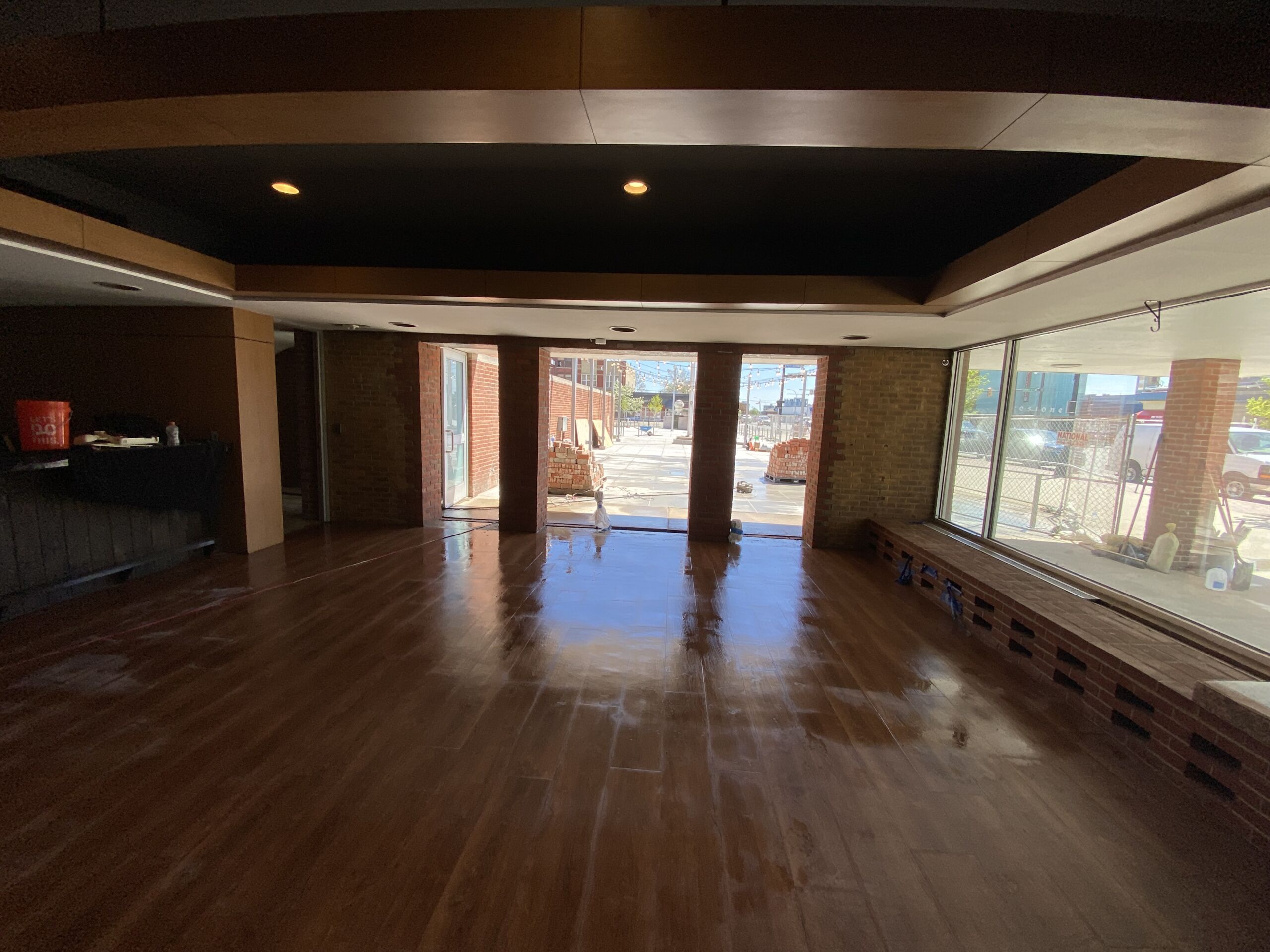 Interior space with dark wood floor, brick columns, and large glass storefront windows showing an outdoor construction scene; ceiling lighting present.