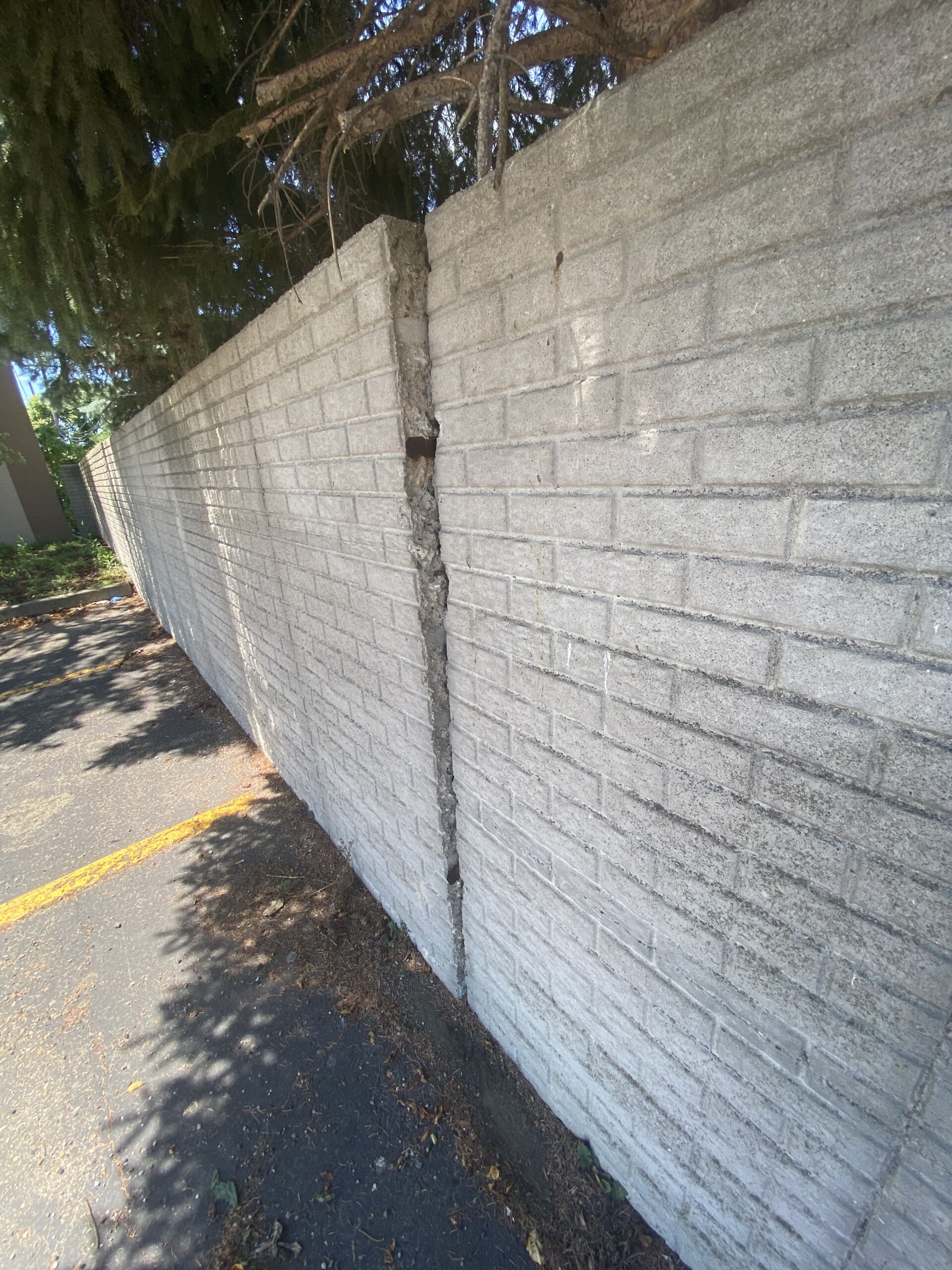 IMG_6954 Concrete block wall with a tall, deep vertical crack running from top to bottom, next to a paved parking area and overhanging tree branches above.