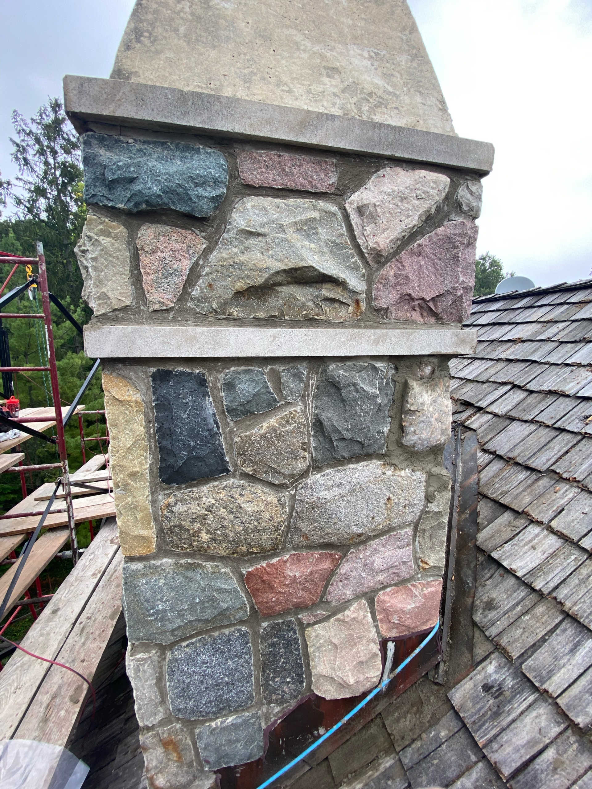 StoneRepair1 Stone chimney built from irregular stones with a flat cap, adjacent to a wooden shingle roof and construction scaffolding nearby.